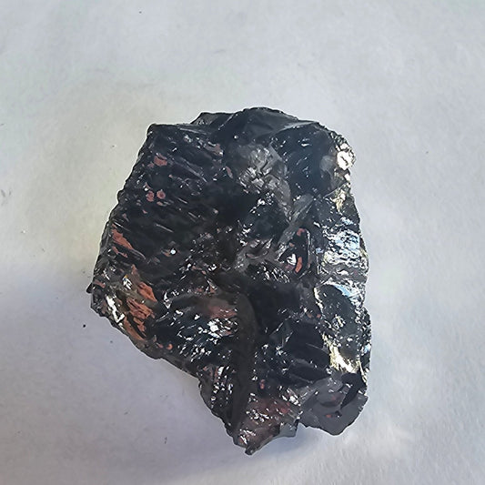 Elite shungite nugget #13, 50g: glossy black faceted chunk with coppery glints, close up on white paper